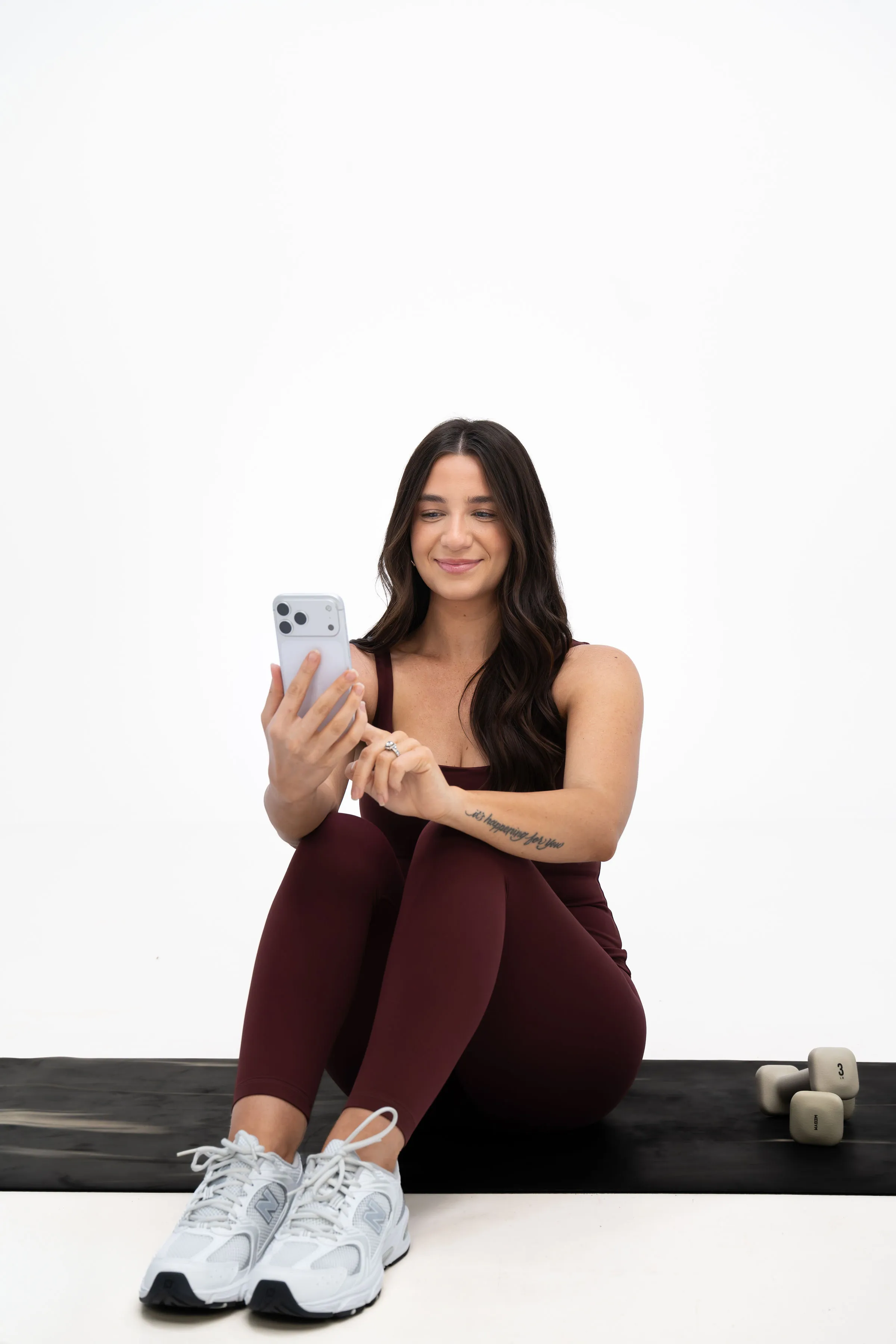 A woman in berry athletic clothing seated on an exercise mat, using her phone.
